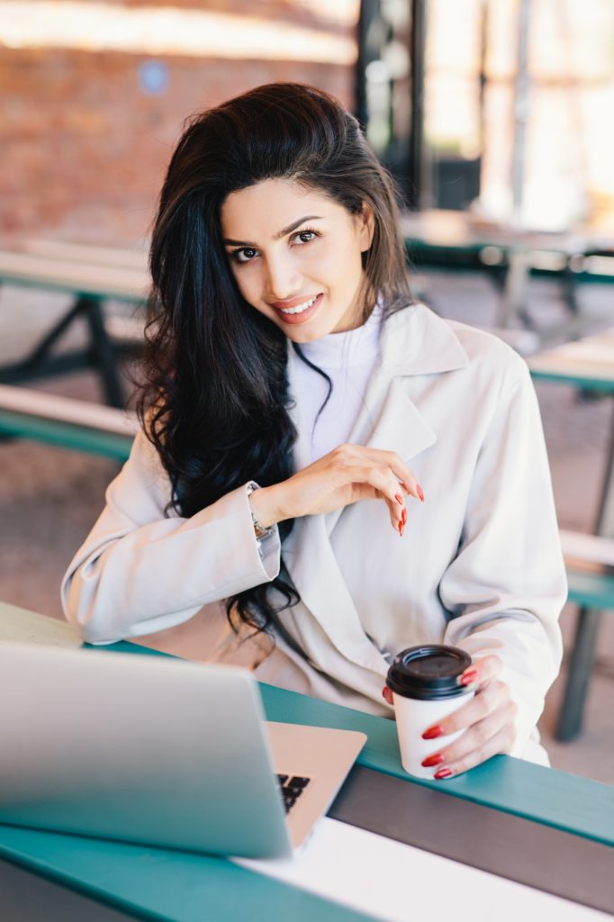 Woman with having bright eyes, full lips and healthy skin wearing white coat resting at cafe